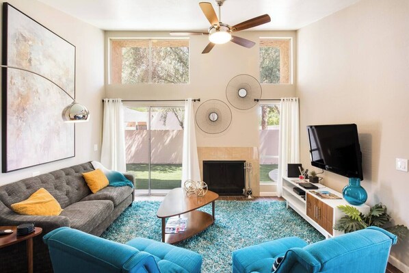 A living room with a sofa, coffee table, and blue chairs. There is a fireplace, a ceiling fan, and a TV mounted on the wall. - Architect-Designed Retreat with a BBQ Patio and Pool (Mesa)