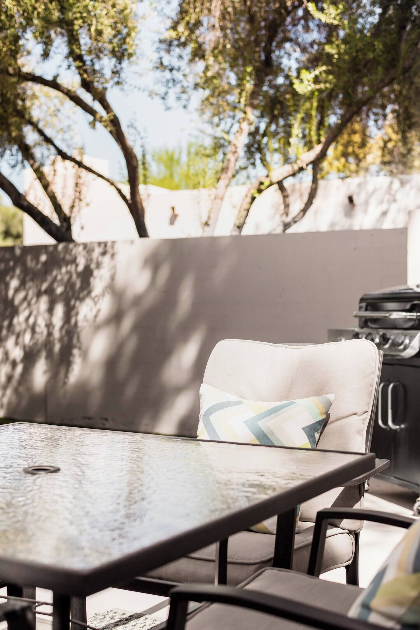 An outdoor seating area with a table and chairs, a grill, and a wall with trees in the background.