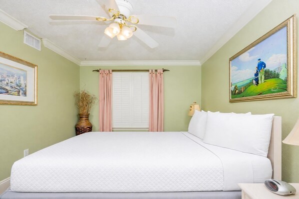 Une chambre à coucher avec un lit, un ventilateur de plafond, une fenêtre avec des rideaux, une table de chevet avec une lampe et un tableau encadré accroché au mur.