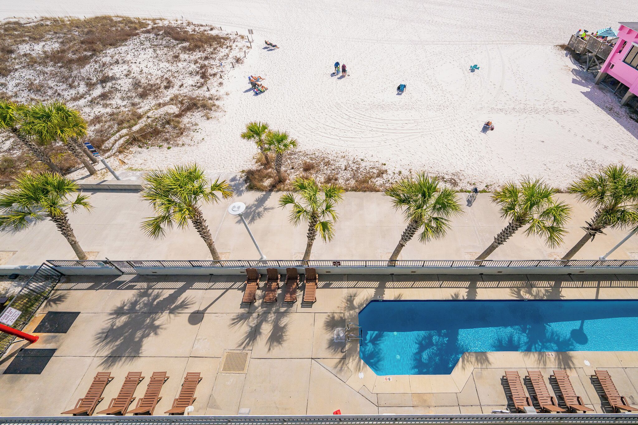 Un espace piscine avec des chaises longues, une piscine et une plage où l’on peut voir des gens et des palmiers.