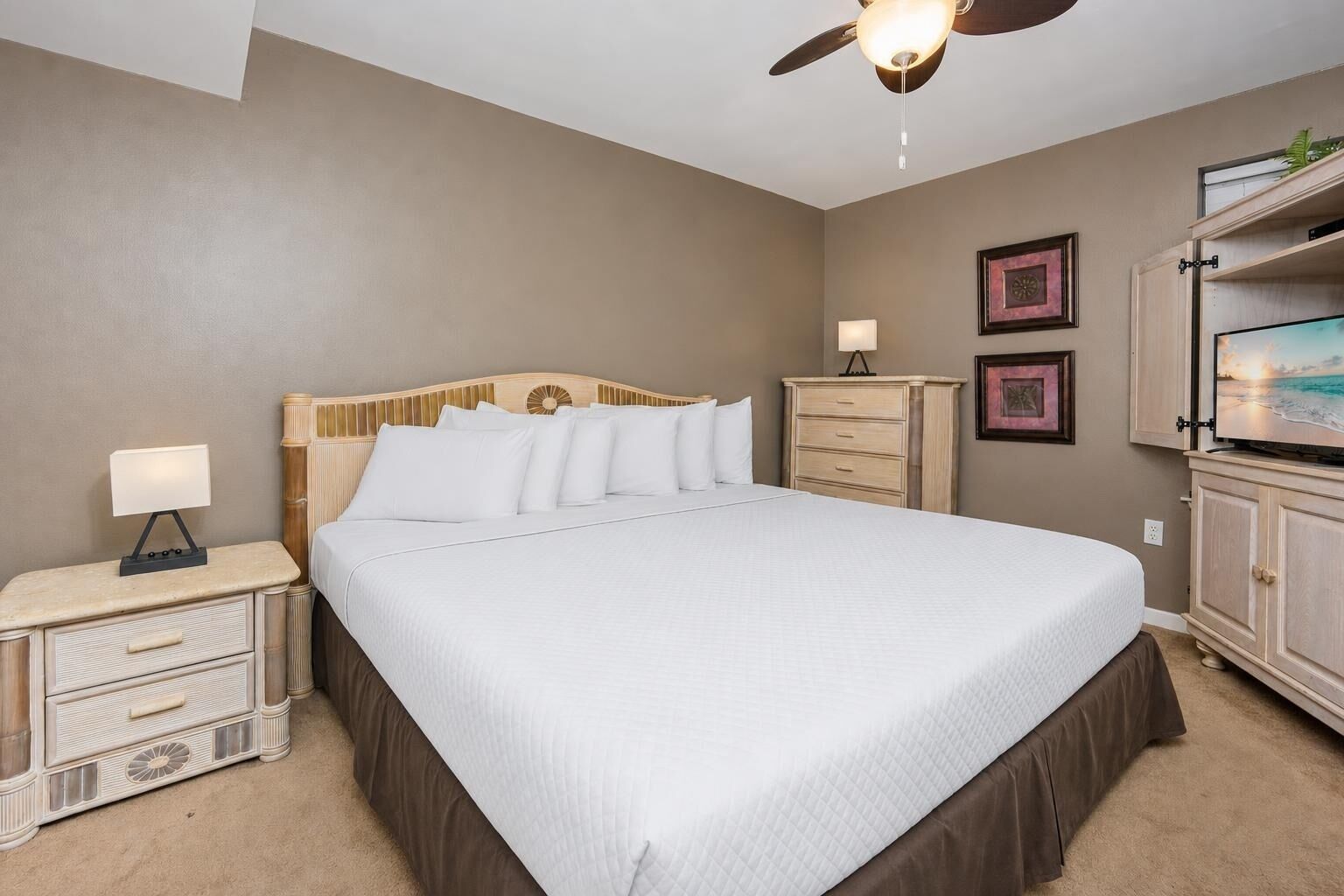 A bedroom with a bed, nightstand, dresser, ceiling fan, and wall-mounted TV.