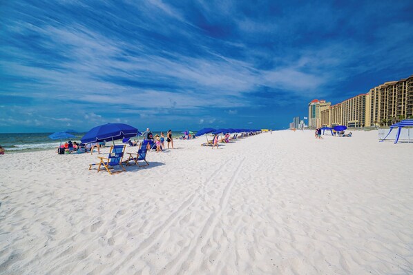 A beach with rows of blue umbrellas and beach chairs, a sandy shore, and a hotel building in the background.