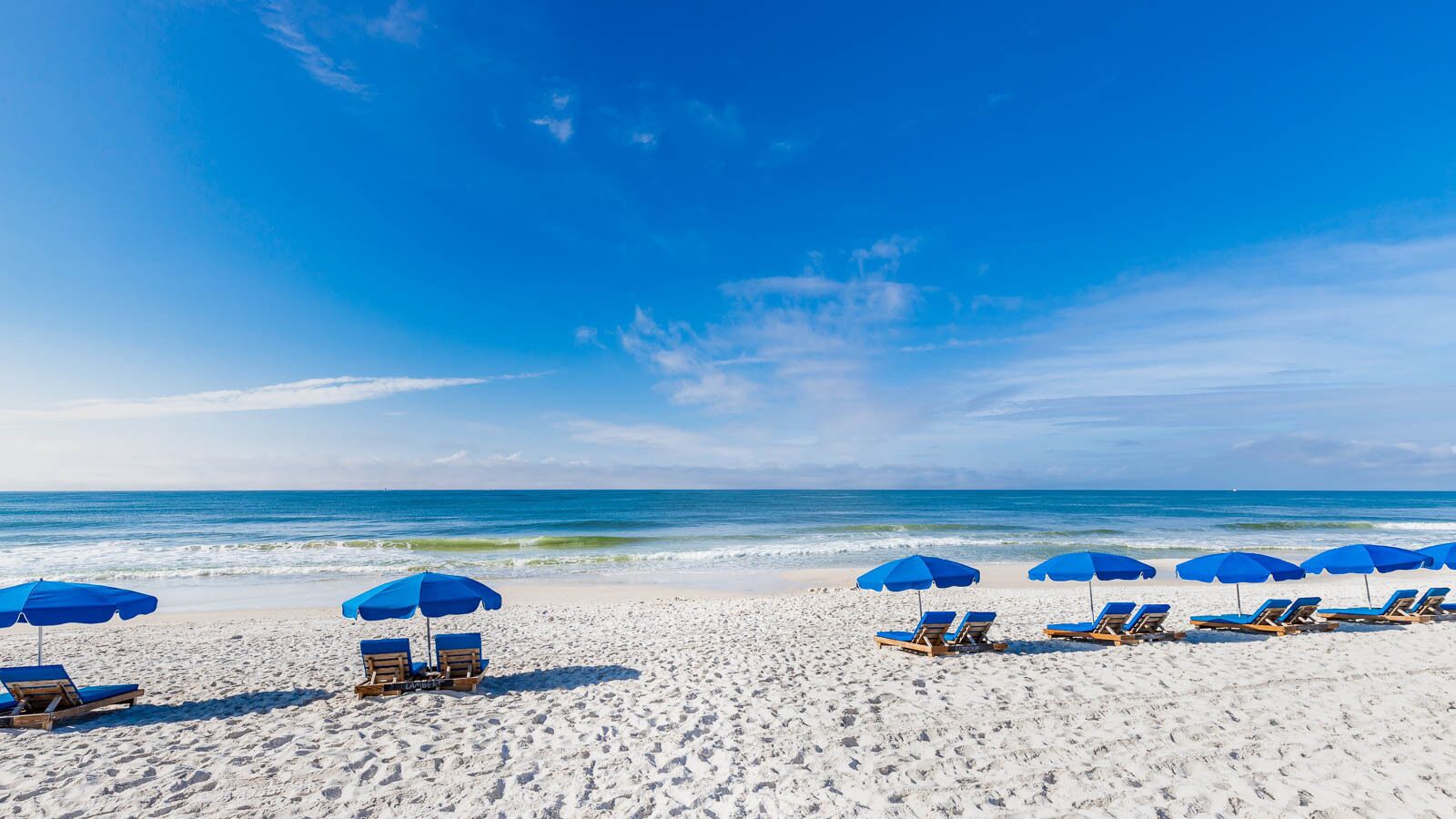 Ein Strand mit weißem Sand und klarem, blauem Wasser. Blaue Sonnenschirme und Holzliegen sind ordentlich in Reihen angeordnet.