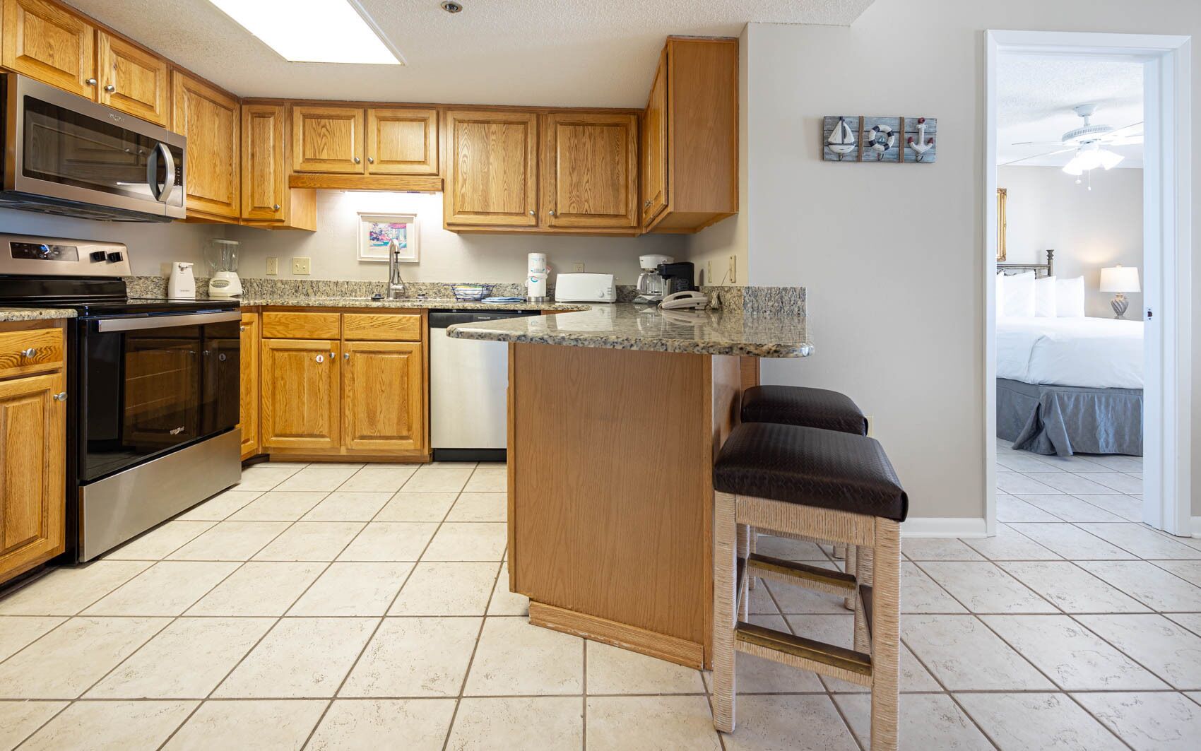 A kitchen with wooden cabinets, a microwave, an oven, and a coffee maker. There is a bar stool and a doorway leading to a bedroom.