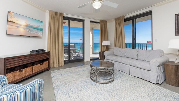 A living room with a sofa, a coffee table, a TV, a ceiling fan, and a view of the ocean through large windows. - Phoenix X 901 - Lovely 3BR Gulf Front End Unit (Orange Beach)