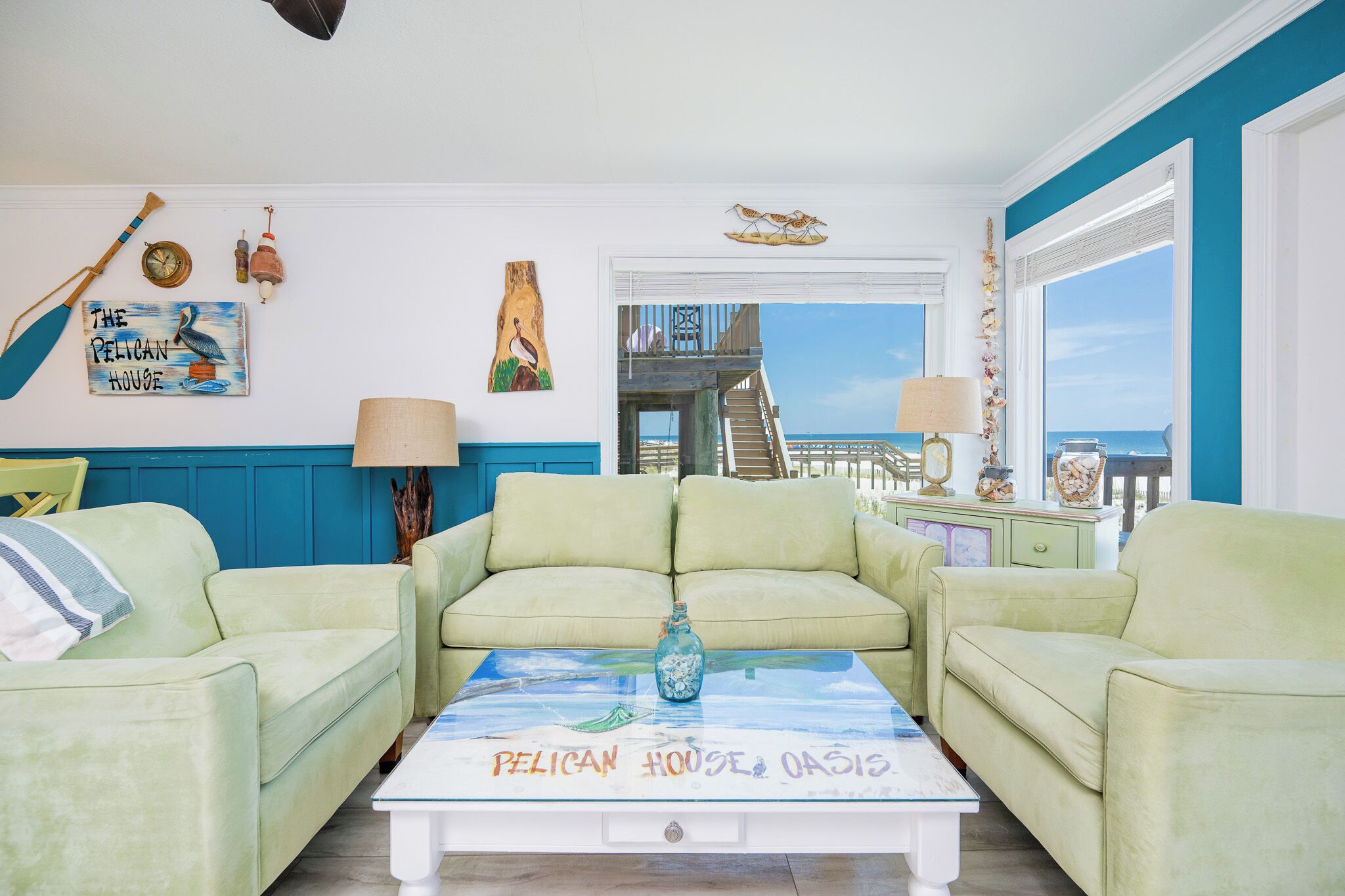 A living room with light green sofas, a coffee table with a 'Pelican House Oasis' artwork, and a view of a balcony with a railing.