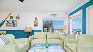 Una sala de estar con sofás verde claro, una mesa de centro con una obra de arte 'Pelican House Oasis', y vistas a un balcón con barandilla.