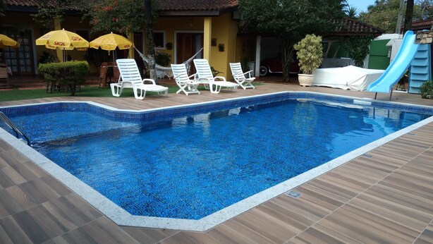 Una piscina rectangular con borde de azulejos, rodeada por una cubierta de madera. Se observan sillas de descanso blancas y sombrillas amarillas con la marca 'SKOL'.