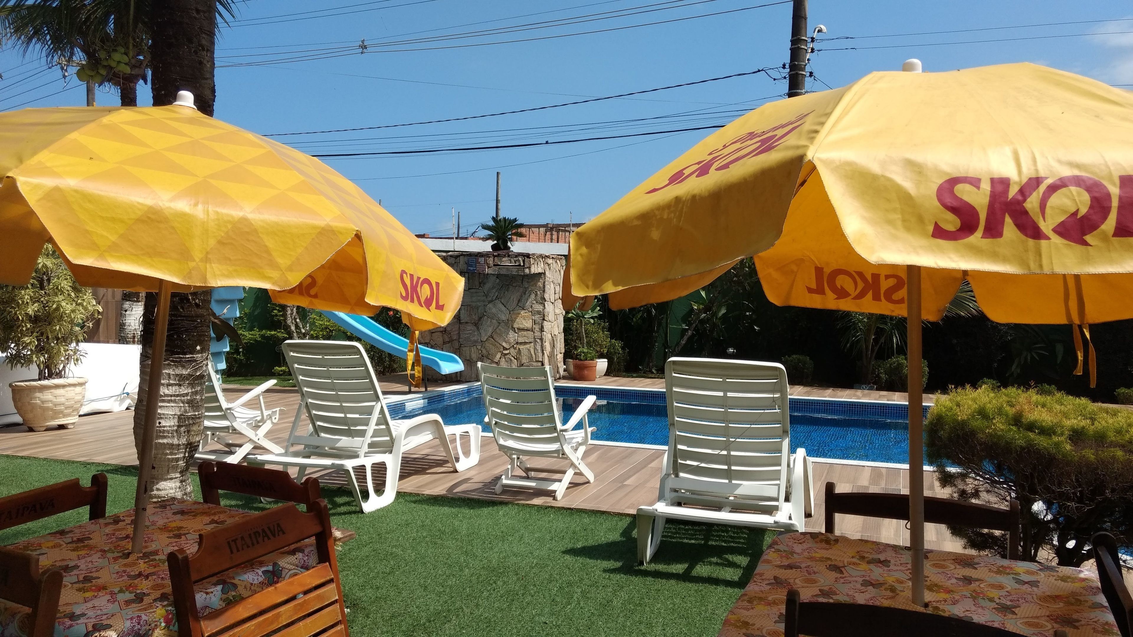 Un área de piscina con sombrillas amarillas que dicen "SKOL", sillones blancos para tomar el sol y una pequeña cascada.