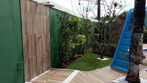 Un espacio exterior moderno con una puerta de madera, un pequeño jardín con diversas plantas, un tobogán azul y un muro de piedra.
