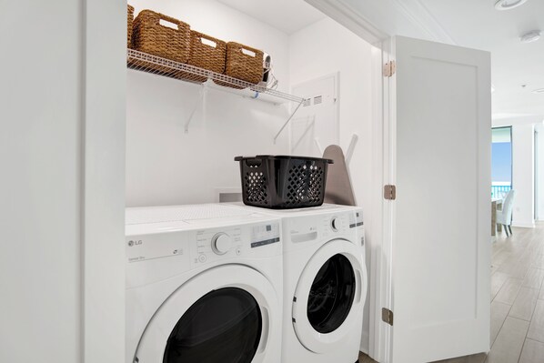 A laundry area with two white front-loading washing machines, a black laundry basket, and wicker baskets on a shelf.