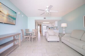 Ein Wohnzimmer mit Deckenventilator, Essbereich mit Stühlen, einer Couch, einem Fernseher und einer Küche im Hintergrund.