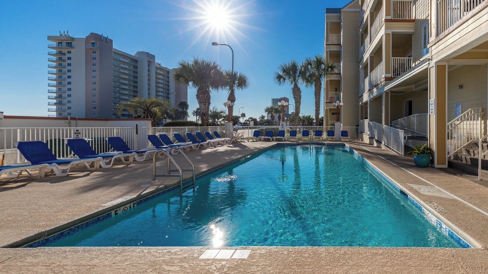 A swimming pool with lounge chairs, a clear sky, and tall buildings in the background.