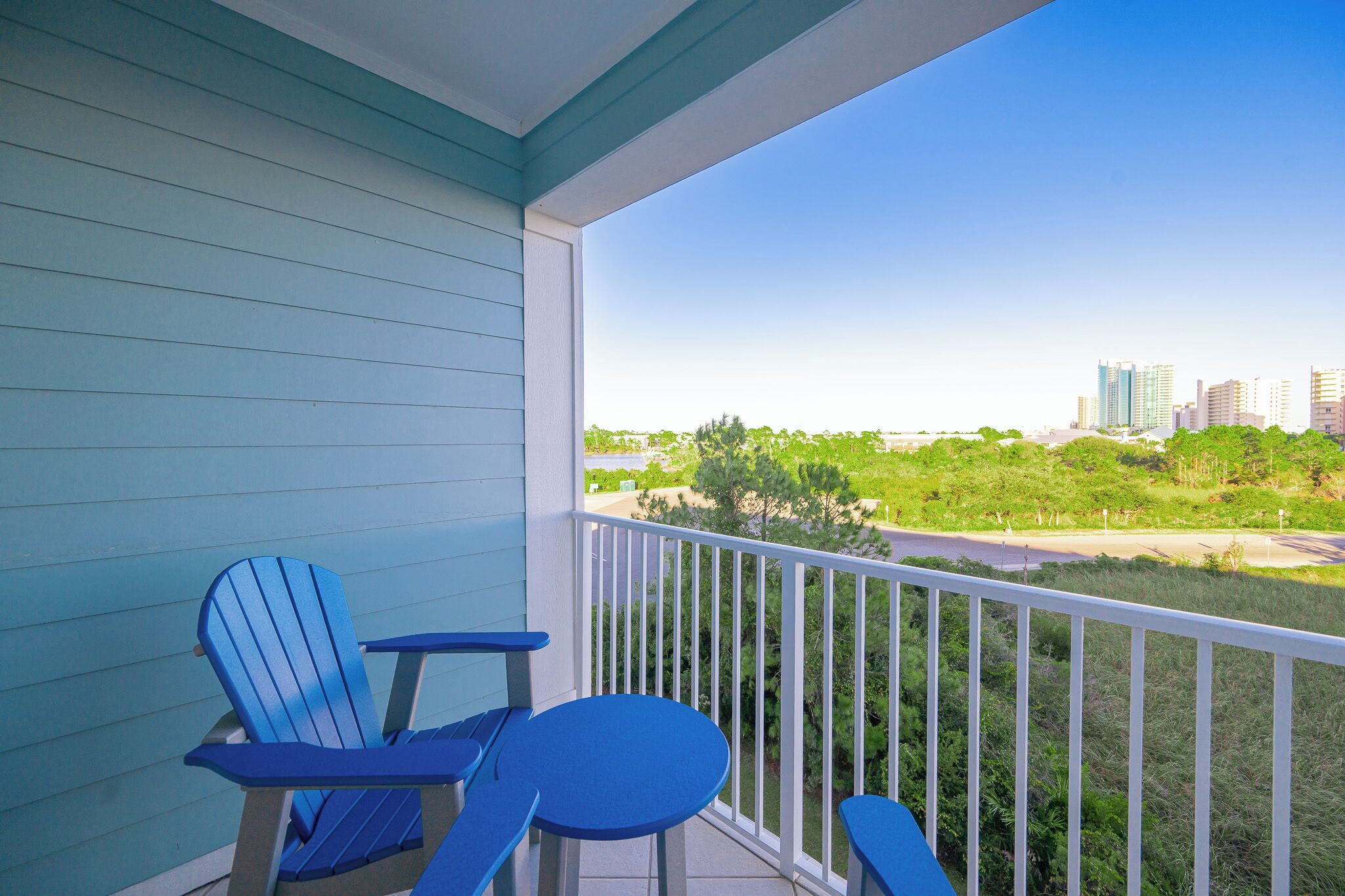 Een balkon met twee blauwe Adirondack stoelen en een tafeltje, met uitzicht op een stadsgezicht met gebouwen en groen.