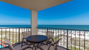 A balcony with a round table and chairs, overlooking a beach with umbrellas and people. - 1 Bedroom @ Phoenix III ~ (Orange Beach)