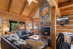 L’intérieur chaleureux d’un chalet en rondins, avec une cheminée en pierre, des poutres en bois au plafond et une grande fenêtre donnant sur une zone boisée.