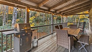 En overdækket terrasse med grill, fletmøbler og udsigt til efterårsfarvede træer.