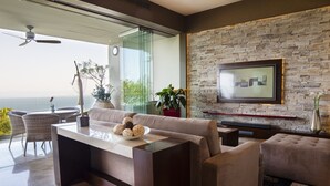 A modern living room with a sofa, a coffee table, and a TV mounted on a stone wall. There is a balcony with a view of the sea.