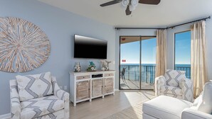 A spacious living room with a large flat-screen TV, a decorative wall piece, and a view of the ocean through a sliding glass door.