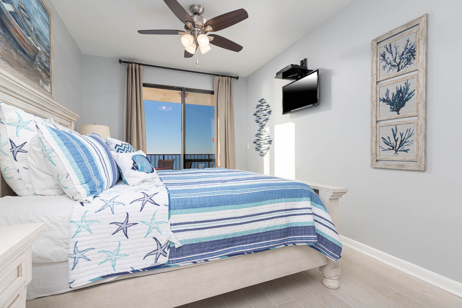 A bedroom with a bed, a ceiling fan, a TV mounted on the wall, and a balcony with curtains.