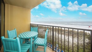 Un balcon avec des chaises turquoise et une table, donnant sur une plage où flotte un drapeau.
