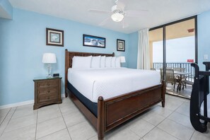 Um quarto com cama, mesas de cabeceira, ventilador no teto e varanda com cadeiras.