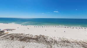 Ein Strand mit Reihen blauer Sonnenschirme und Liegen, ein sandiger Uferstreifen und das Meer im Hintergrund.