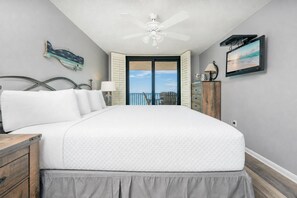Une chambre à coucher comprenant un lit, une table de chevet, une commode, un ventilateur de plafond, un téléviseur et une vue sur l’extérieur.
