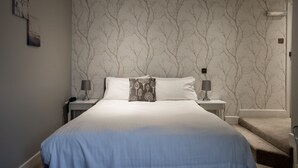 A bedroom with a large bed, two bedside tables, a nightstand, and a wall with a tree pattern. - Ormonde House Hotel (Lyndhurst)