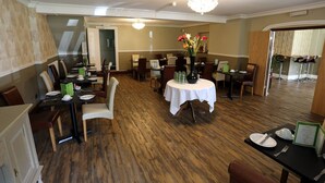 A dining area with wooden flooring, round tables, and chairs. There are flower vases on the tables and a chandelier hanging from the ceiling. - Ormonde House Hotel (Lyndhurst)