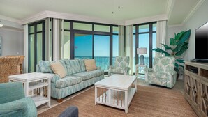 Un salón amplio con un sofá grande, dos sillones, una mesa de centro y un televisor sobre un mueble de madera. La habitación tiene ventanas grandes con vista al océano.