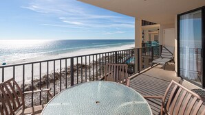 A balcony with a glass table and chairs, overlooking a beach and ocean.