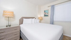 A neatly made bed with white bedding, a wicker nightstand with drawers, a lamp with a chevron pattern, and a wall-mounted picture.