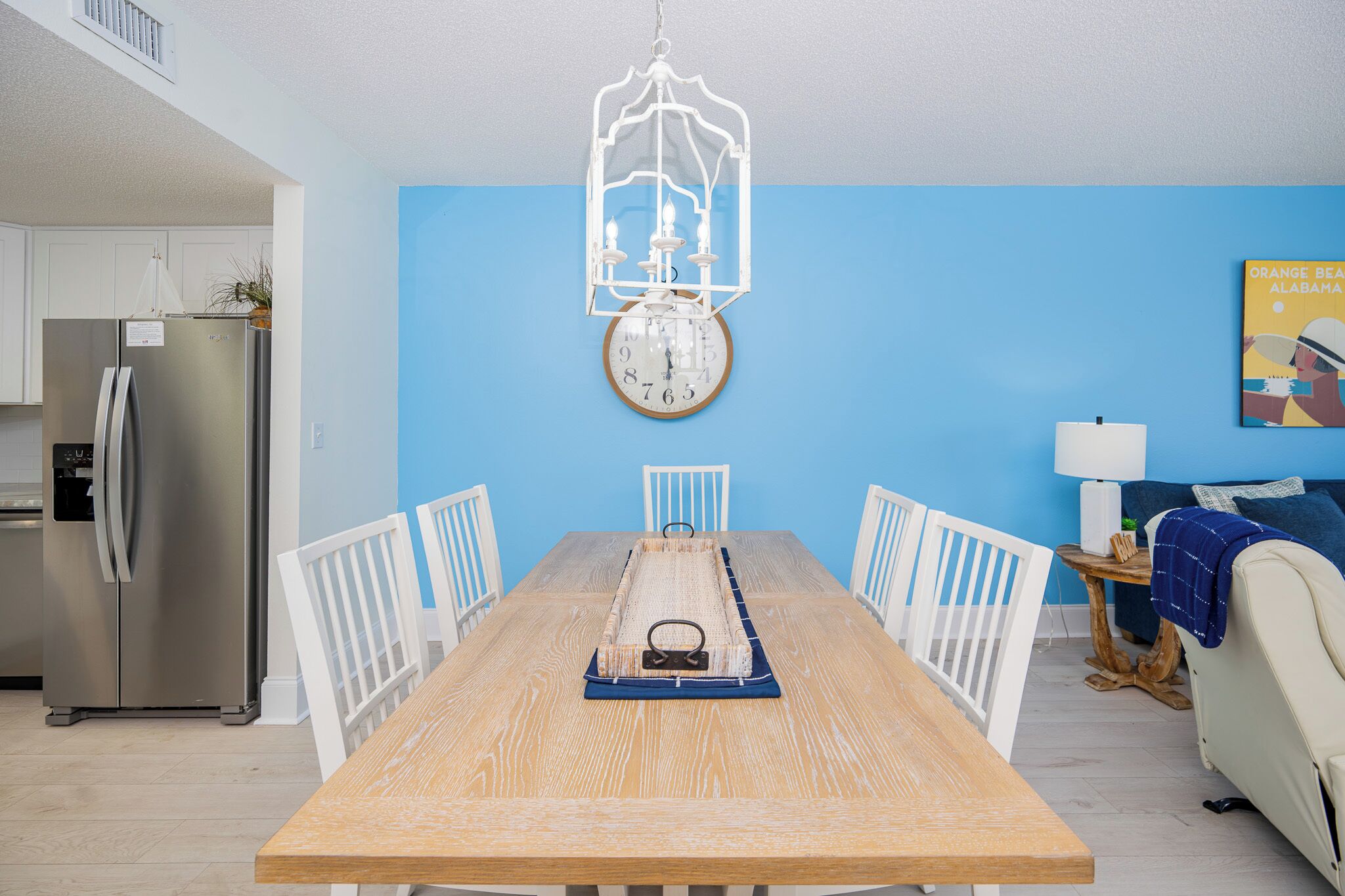 A dining room with a wooden table, white chairs, a hanging light fixture, and a clock on the wall.