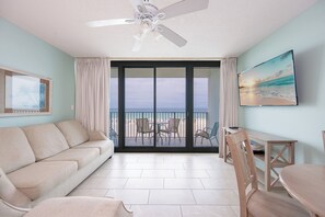 Ein Zimmer mit einer Couch, einem Esstisch, Stühlen, einem Deckenventilator, einem Gemälde und Blick auf den Strand durch eine Schiebetür.
