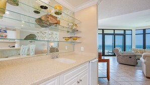 A modern kitchen with a glass display shelf, a sink, and a view of the living area with a balcony.
