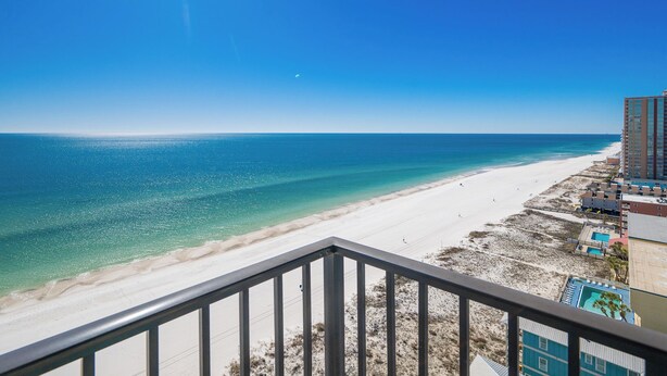 A beach view from a balcony with a clear sky, a sandy beach, and a building with a pool.