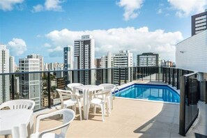 Una terraza en la azotea con piscina, sillas de plástico blanco y un horizonte urbano al fondo.
