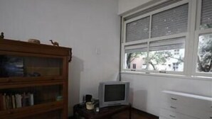 Una habitación con un armario de madera, un televisor sobre una mesita y una ventana con persianas.