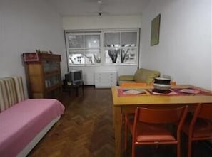 A small room with a bed, a table with chairs, a radiator, a window, and a television.