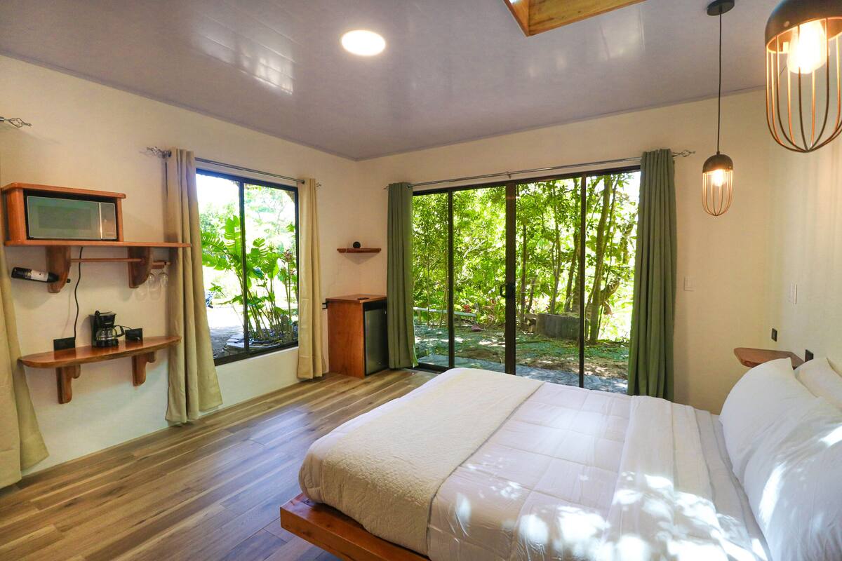 Un dormitorio con una cama grande, suelo de madera, un televisor en un estante y vistas a la vegetación exterior.