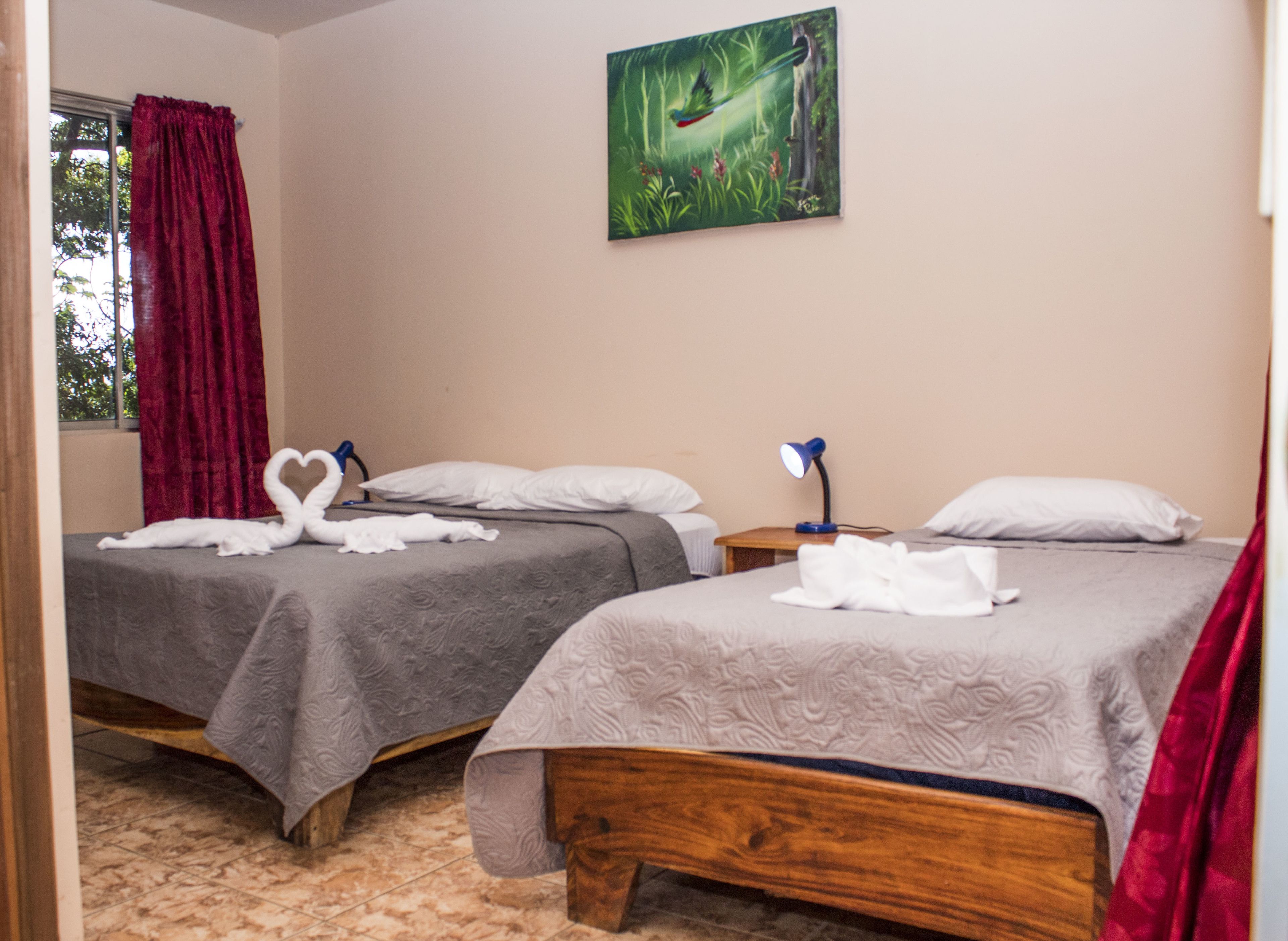 Ein Hotelzimmer mit zwei Betten, jedes mit einem herzförmigen Handtuch, einem Gemälde von einem Vogel im Wald und einem Fenster mit roten Vorhängen.