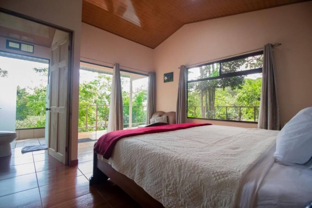 Un dormitorio con una cama grande, techo de madera y balcón con vistas a la vegetación.