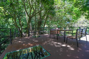 Un área de estar al aire libre con mesas y bancos, rodeada de árboles y una barandilla de metal.