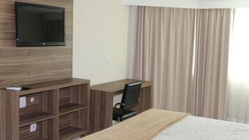 Una habitación de hotel con una cama, un escritorio con silla, un televisor montado en la pared y una ventana con cortinas.