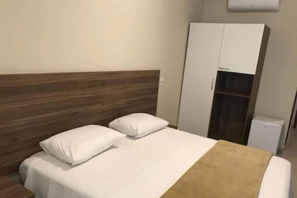 A hotel room with a wooden headboard, a bed with white bedding and a brown throw, a wardrobe, and a small white appliance.