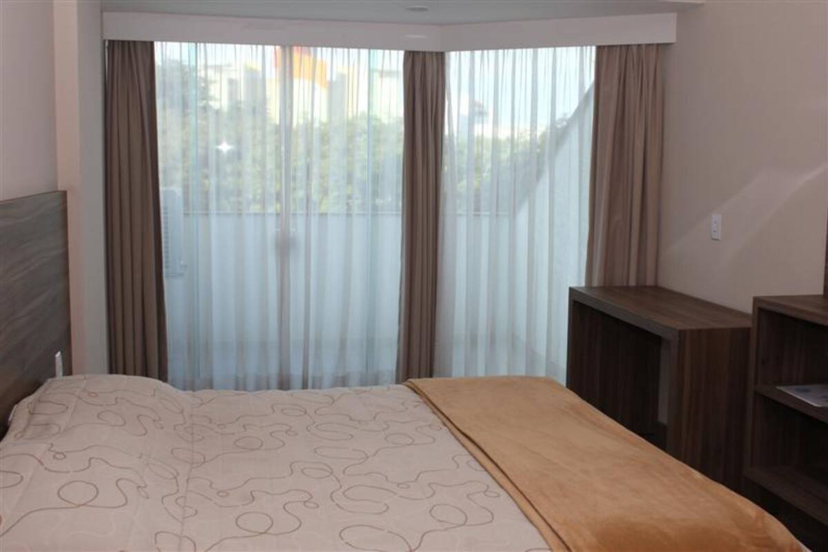 A hotel room with a bed, a wooden nightstand, and a large window with sheer curtains.