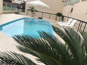 Una piscina con una sombrilla blanca, rodeada de palmeras y sillas de descanso blancas.