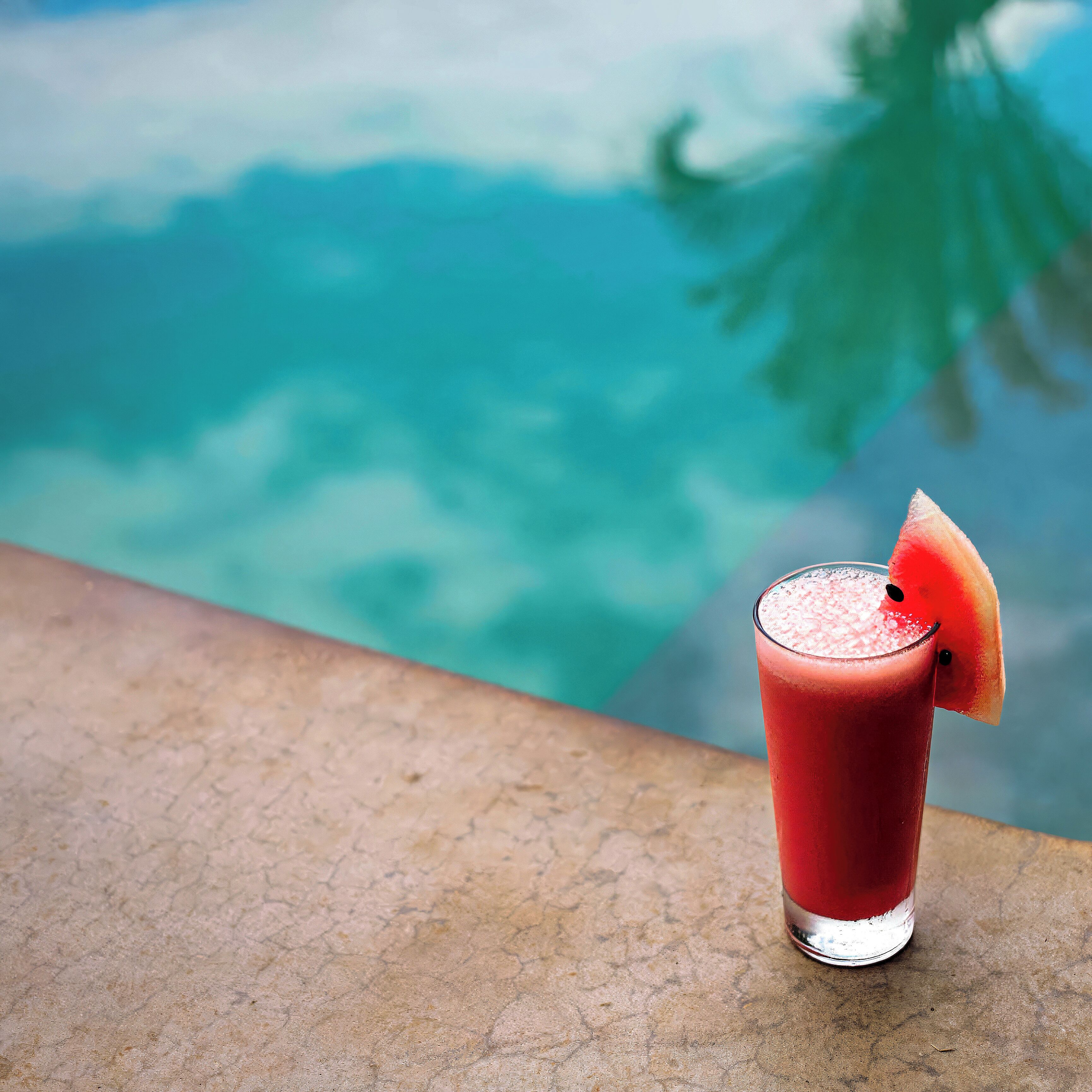 A glass of red beverage with a slice of watermelon on the rim, placed on a textured surface with a blurred background of a body of water and greenery.