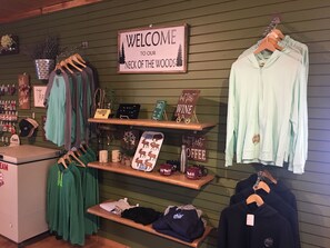 A clothing store with a 'Welcome to our Neck of the Woods' sign, shelves with clothing, and signs for 'But First Wine' and 'But First Coffee'.